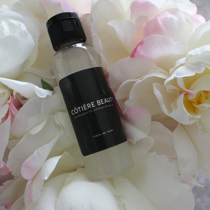 COTIÈRE BEAUTY Nourishing Hair Oil bottle displayed on a bed of white and pink peonies, showcasing luxury beauty product in a soft floral setting